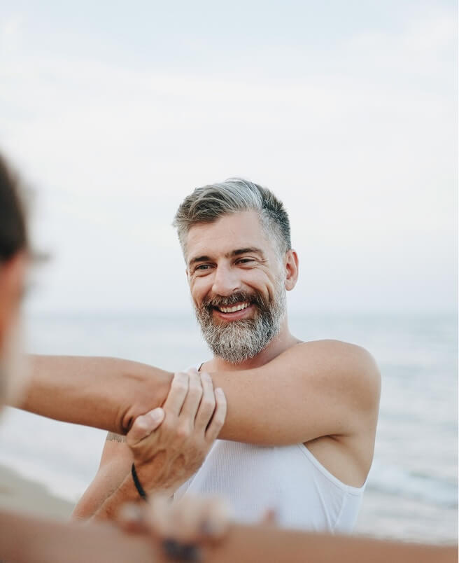 A man stretching his arm while smiling at another person on the beach